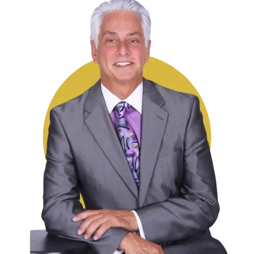 A man with gray hair wearing a gray suit, purple shirt, and patterned tie is seated and smiling, with a yellow circle in the background.