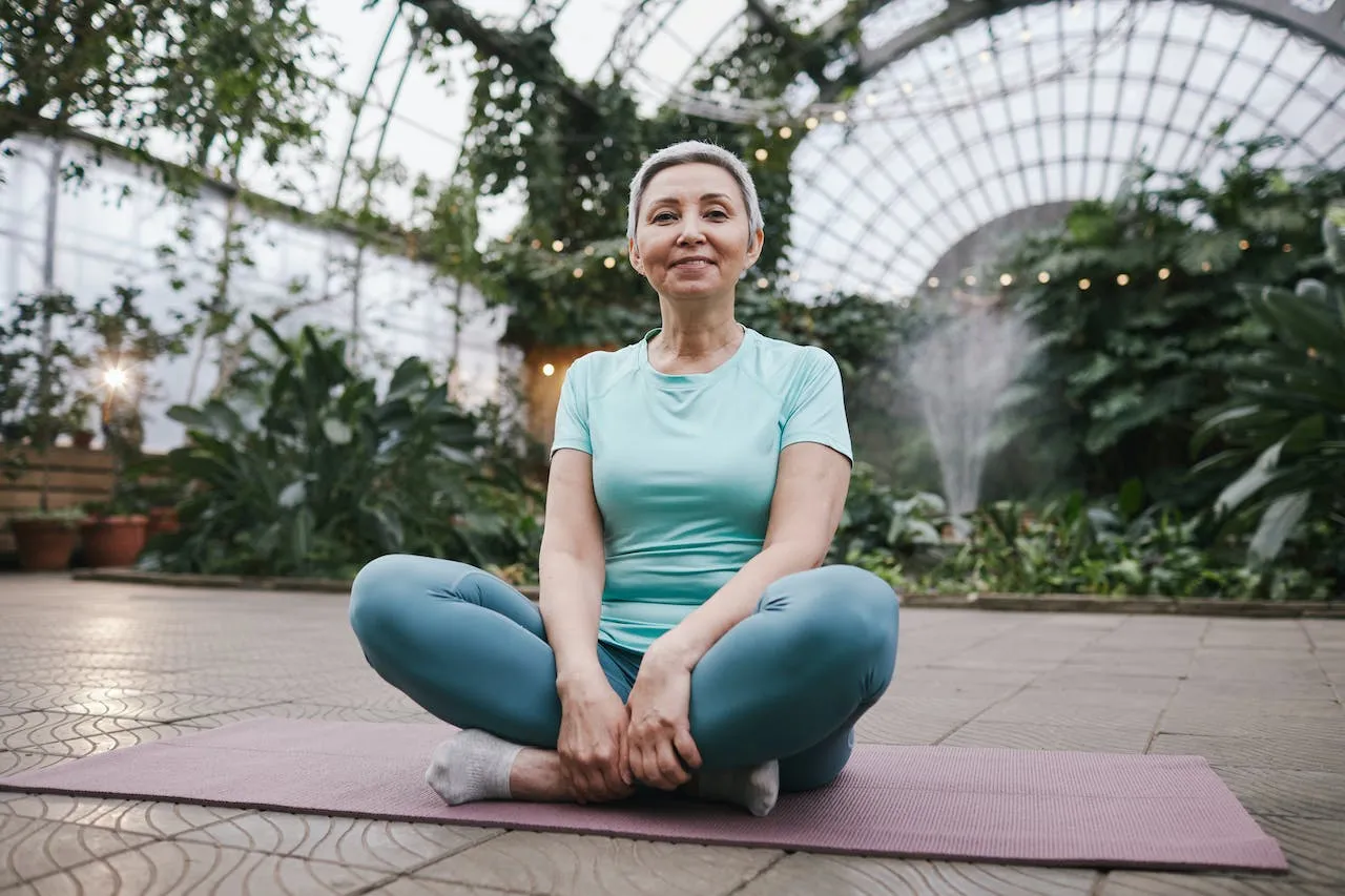 An older woman in athletic wear sits cross-legged on a yoga mat inside a greenhouse filled with plants.