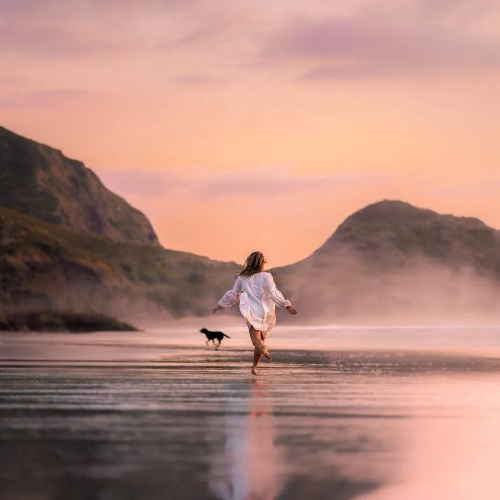 A woman in a white dress runs barefoot on a beach at sunset, with a dog nearby and hills in the background.