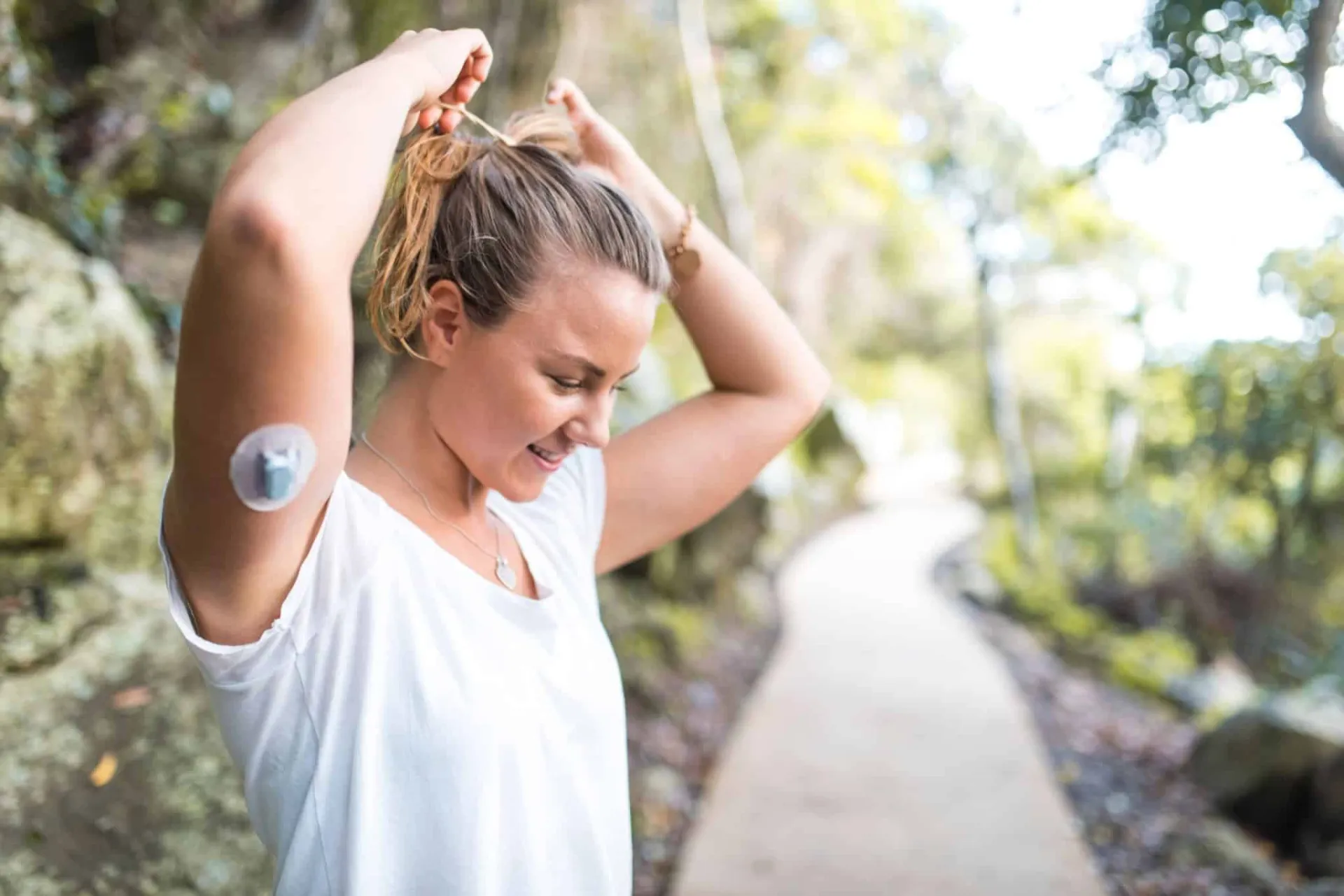 A woman in a white shirt ties her hair back outdoors, with a glucose monitor on her upper arm.